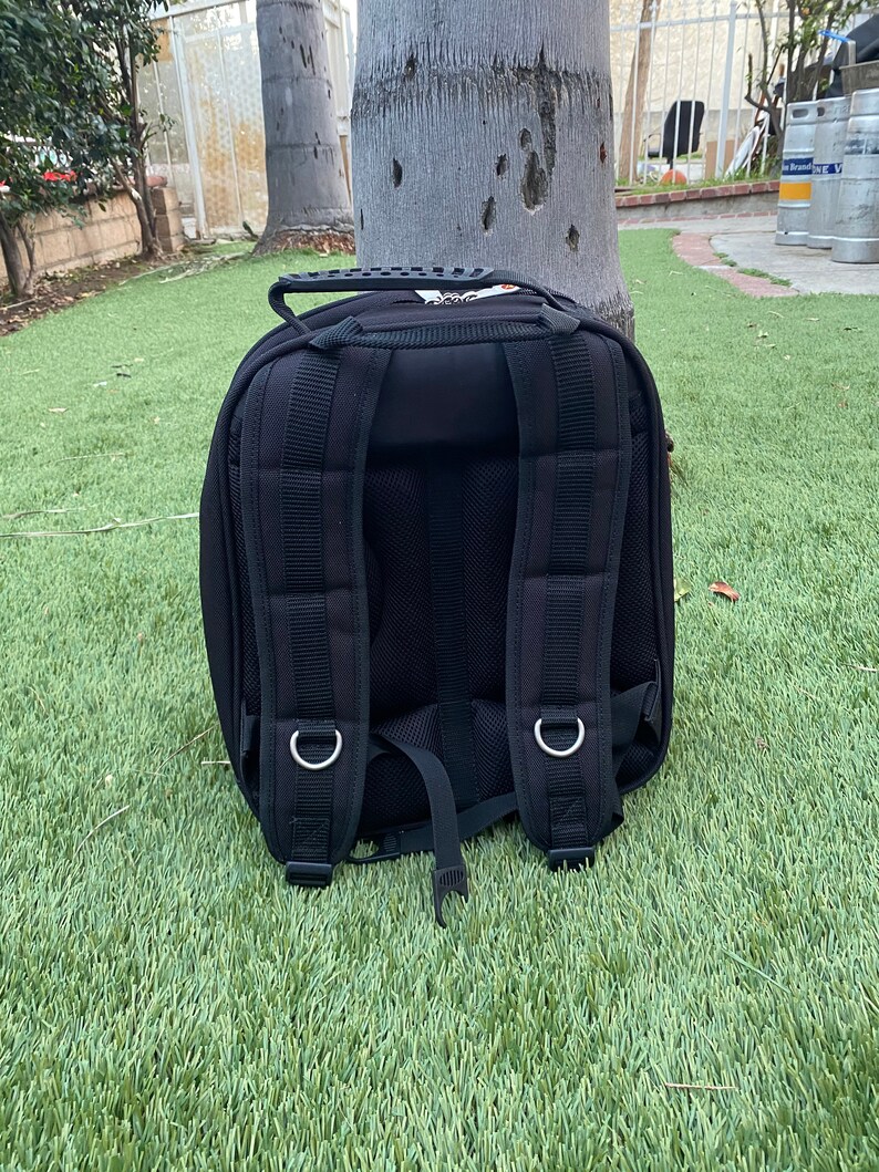 May include: Black backpack with padded shoulder straps, a top handle, and multiple compartments. The backpack is made of a durable material and features silver metal rings for attaching accessories. It is sitting on green grass.