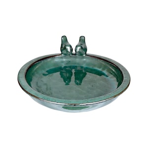 Può includere: Un bagno per uccelli circolare in ceramica color verde acqua, caratterizzato da una finitura liscia e lucida con dettagli marrone scuro sul bordo. Due piccole statuette di uccelli stilizzate sono fissate sul bordo, rivolte verso il centro.
