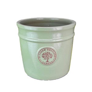 Pode incluir: Vaso de jardim de cerâmica verde claro com um emblema redondo "Blossom Orchard Garden". O vaso tem uma abertura larga e uma superfície texturizada. Ideal para plantar flores ou ervas.