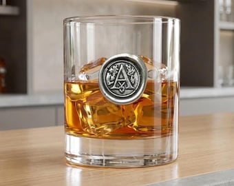 Może przedstawiać: Szkło do whisky wypełnione lodem i bursztynowym płynem, prawdopodobnie whisky. Srebrny emblemat z literą "A" jest przymocowany do szkła. Szkło stoi na drewnianej powierzchni.