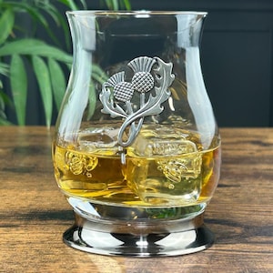 May include: A clear glass whisky tumbler with a silver thistle design. The glass is filled with amber liquid and ice cubes.