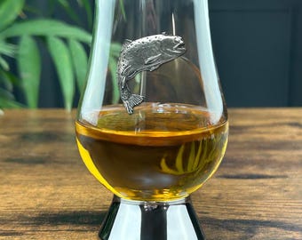 Może przedstawiać: Szklanka do whisky z przezroczystego szkła z srebrną podstawą. Po bokach szklanki znajduje się figurka pstrąga z cyny.
