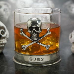 May include: A clear glass tumbler with a pewter base featuring a skull and crossbones design. The glass is filled with amber liquid and ice cubes. The pewter base is engraved with the letters "EPC".
