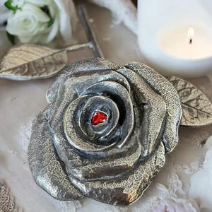 May include: A silver rose with a red heart in the centre. The rose is a decorative object with a textured surface.