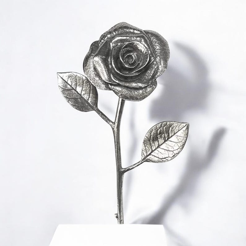 May include: A silver metal rose with two leaves on a stem. The rose is detailed and realistic.