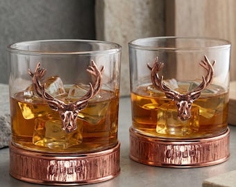 May include: Two clear whiskey glasses with copper-colored deer head embellishments. The glasses are filled with amber liquid and ice cubes. The base of each glass is copper-colored with etched details. A sophisticated barware set.