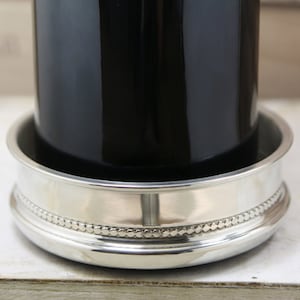 May include: A silver wine coaster with a beaded edge, holding a dark glass bottle. The coaster's rim features a decorative beaded pattern. The coaster has a reflective surface.