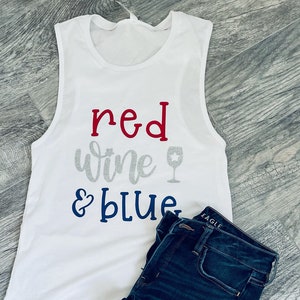 May include: White tank top with the text "red wine & blue" in red, silver, and blue lettering. A wine glass icon is next to the word "wine". The tank top is laying on a wooden surface with a pair of blue jeans with rips.