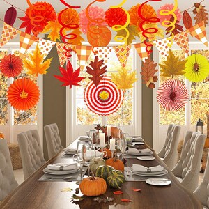 May include: A Thanksgiving table setting with fall-themed decorations. The table is set with white plates, silverware, and glasses. There are fall-colored paper fans, leaves, and a banner hanging above the table. There are also pumpkins and gourds on the table.