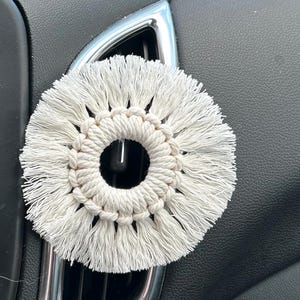 Może przedstawiać: Kremowy odświeżacz powietrza do samochodu z makramy o okrągłym kształcie. Odświeżacz powietrza wykonany jest z tkanej bawełny i posiada centralny otwór. Umieszczony jest na kratce wentylacyjnej samochodu, na tle czarnej deski rozdzielczej.