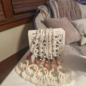 Pode incluir: Uma cole&ccedil;&atilde;o de cinco penduradores de parede em macram&eacute;. Cada um apresenta um anel de madeira com uma base com franjas, conectado por uma corda branca a contas de madeira. As contas s&atilde;o castanhas, vermelhas e estampadas. Os penduradores s&atilde;o exibidos em frente a uma caixa decorativa branca.