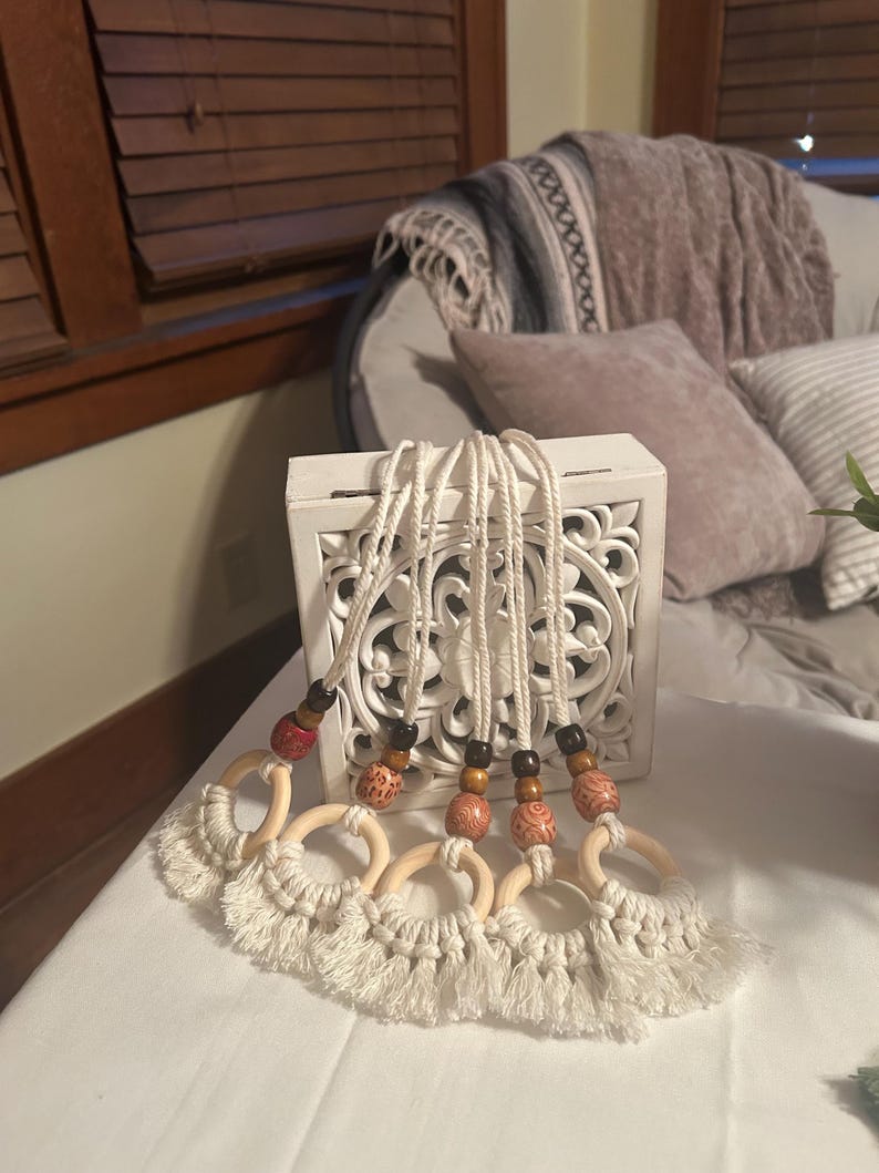 Pode incluir: Uma cole&ccedil;&atilde;o de cinco penduradores de parede em macram&eacute;. Cada um apresenta um anel de madeira com uma base com franjas, conectado por uma corda branca a contas de madeira. As contas s&atilde;o castanhas, vermelhas e estampadas. Os penduradores s&atilde;o exibidos em frente a uma caixa decorativa branca.