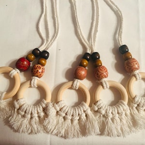 Pode incluir: Cinco amuletos de carro em macram&eacute; com an&eacute;is de madeira e contas decorativas. Cada amuleto apresenta um anel de madeira natural com uma franja de macram&eacute; branca. As contas s&atilde;o uma mistura de designs castanhos, vermelhos e estampados, suspensos por um cord&atilde;o branco.