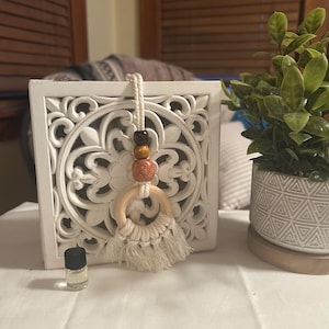 Op de afbeelding: Witte, gebeeldhouwde houten wanddecoratie met een macrame etherische olie diffuser. De diffuser heeft een houten ring, crèmekleurige franjes en houten kralen. Een klein glazen flesje etherische olie staat voor de decoratie, naast een kleine potplant.