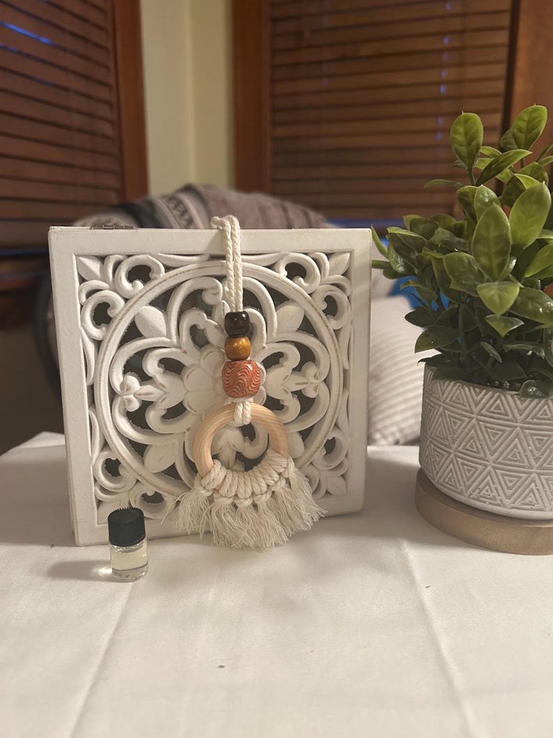 Pode incluir: Decora&ccedil;&atilde;o de parede de madeira entalhada branca com um purificador de ar para carro de macram&ecirc; e contas de madeira pendurado na frente. Uma pequena garrafa de vidro com tampa preta est&aacute; sobre a mesa. Uma pequena planta em vaso est&aacute; &agrave; direita.