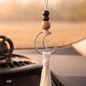 May include: A car charm featuring a silver crescent moon pendant with a cream-colored tassel. The charm is adorned with wooden beads and hangs from a white cord, adding a touch of bohemian style to any vehicle.