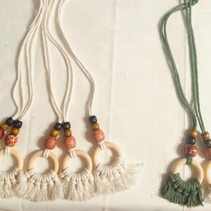 Pode incluir: Enfeites de anel de macram&eacute; com contas de madeira e franjas. Os enfeites apresentam an&eacute;is de madeira natural, franjas de macram&eacute; creme e contas decorativas em vermelho, castanho e laranja. Um conjunto tem franjas de macram&eacute; verdes.