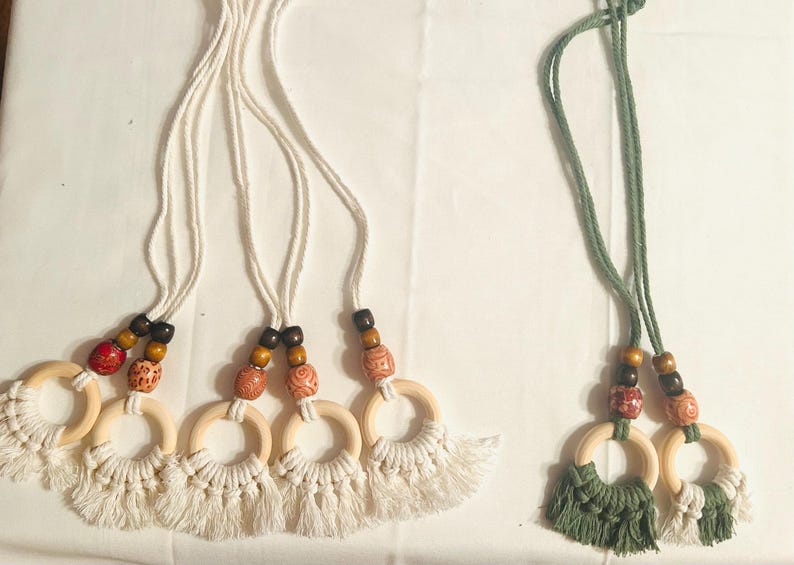 Pode incluir: Enfeites de anel de macram&eacute; com contas de madeira e franjas. Os enfeites apresentam an&eacute;is de madeira natural, franjas de macram&eacute; creme e contas decorativas em vermelho, castanho e laranja. Um conjunto tem franjas de macram&eacute; verdes.