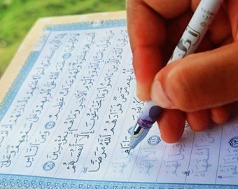 May include: A hand holding a pen and writing in a book with Arabic text. The book has a blue and white border.