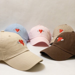 Puede incluir: Cuatro gorras de béisbol de diferentes colores con un diseño de corazón rojo bordado. El corazón está roto por la mitad y sostenido por dos manos. Las gorras son azul claro, rosa, beige y marrón.