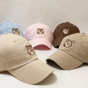 May include: Five baseball caps in different colors, including light blue, pink, brown, and two shades of beige. Each cap features an embroidered cartoon bear with brown fur and pink eyes.