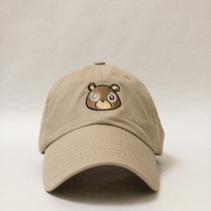 May include: A beige baseball cap with a brown bear embroidered on the front. The bear has red eyes with white pupils.