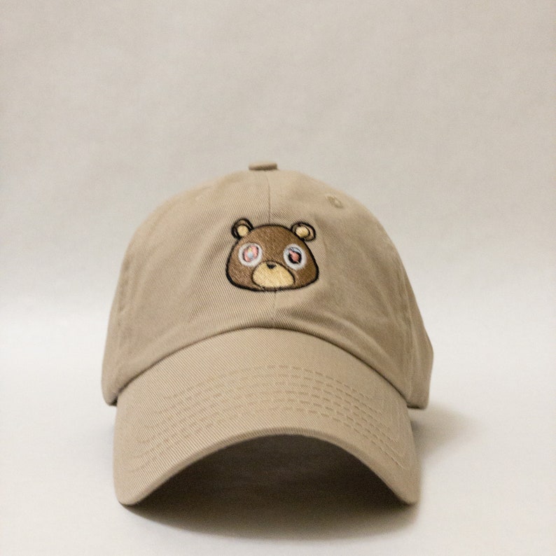 May include: A beige baseball cap with a brown bear embroidered on the front. The bear has red eyes with white pupils.