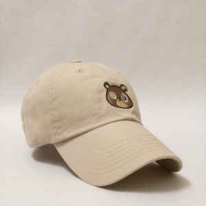 May include: A beige baseball cap with a brown bear embroidered on the front. The bear has one pink eye and one brown eye.