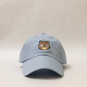 May include: A light blue baseball cap with a brown bear embroidered on the front. The bear has pink eyes and a brown nose.
