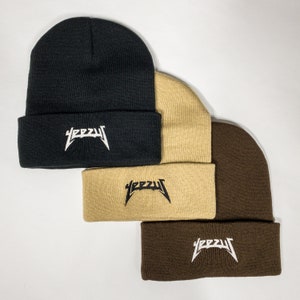 May include: Three beanie hats stacked on top of each other. The top hat is black, the middle hat is beige, and the bottom hat is brown. All three hats have the word "YEEZY" embroidered on the front in white letters.