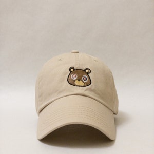 May include: A beige baseball cap with a brown bear embroidered on the front. The bear has pink eyes and a black nose.