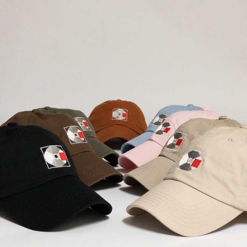K&ouml;nnte beinhalten: Sechs Baseballkappen in verschiedenen Farben mit einem gestickten Schallplatten-Design auf der Vorderseite. Die Kappen sind in einem Halbkreis auf einer wei&szlig;en Oberfl&auml;che angeordnet.