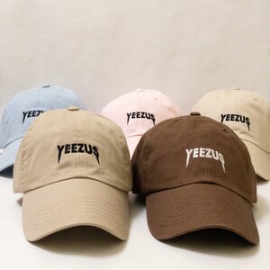 K&ouml;nnte beinhalten: F&uuml;nf Baseballkappen in verschiedenen Farben, darunter hellblau, rosa, beige und braun. Jede Kappe tr&auml;gt den Schriftzug "YEEZUS" in Schwarz oder Wei&szlig; auf der Vorderseite.