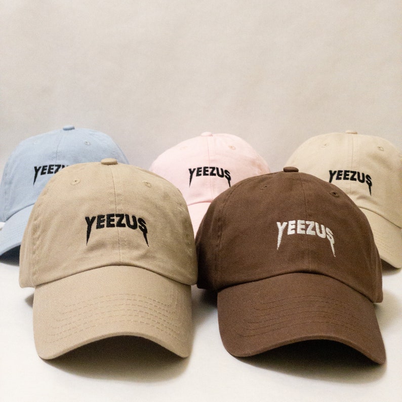 K&ouml;nnte beinhalten: F&uuml;nf Baseballkappen in verschiedenen Farben, darunter hellblau, rosa, beige und braun. Jede Kappe tr&auml;gt den Schriftzug "YEEZUS" in Schwarz oder Wei&szlig; auf der Vorderseite.