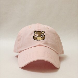 May include: A pink baseball cap with a brown bear embroidered on the front. The bear has brown eyes with a swirl pattern.