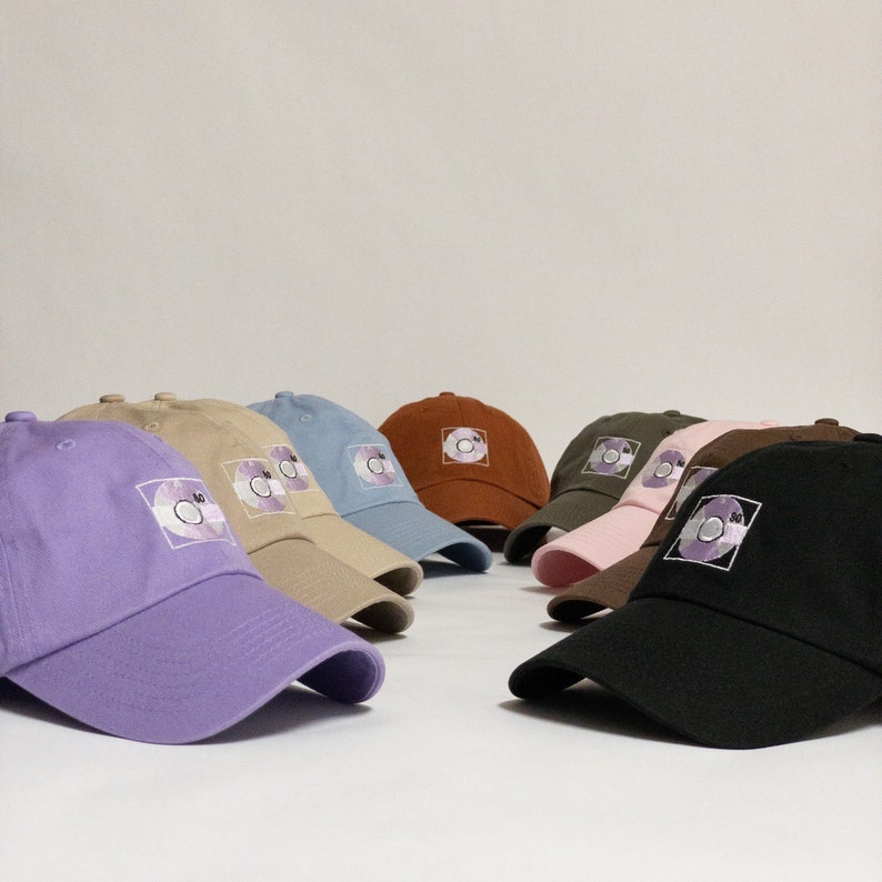 May include: A collection of ten baseball caps in various colours, including purple, pink, brown, blue, and beige. Each cap features a circular design with a purple and white pattern.