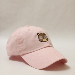 May include: A pink baseball cap with a brown bear embroidered on the front. The bear has brown eyes and a brown nose.