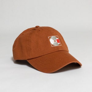 K&ouml;nnte beinhalten: Eine braune Baseballkappe mit einem wei&szlig;en und roten gestickten Logo einer Schallplatte auf der Vorderseite.
