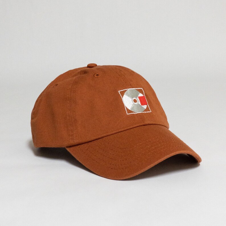 K&ouml;nnte beinhalten: Eine braune Baseballkappe mit einem wei&szlig;en und roten gestickten Logo einer Schallplatte auf der Vorderseite.
