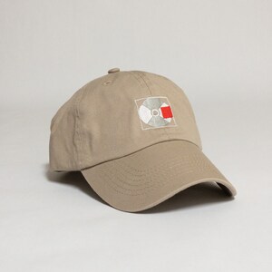 K&ouml;nnte beinhalten: Eine beige Baseballkappe mit einem gestickten wei&szlig;en und roten Schallplattenlogo. Das Logo ist eine stilisierte Schallplatte mit einem roten Quadrat in der oberen rechten Ecke.