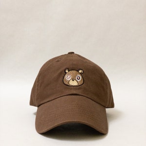 May include: A brown baseball cap with an embroidered brown bear with pink eyes. The bear has a brown nose and mouth.