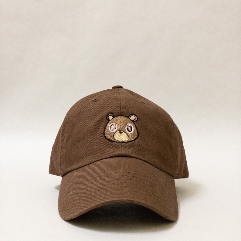 May include: A brown baseball cap with an embroidered brown bear with pink eyes. The bear has a brown nose and mouth.