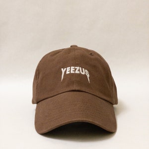 K&ouml;nnte beinhalten: Eine braune Baseballkappe mit einem wei&szlig;en "YEEZUS"-Logo, das auf der Vorderseite gestickt ist.