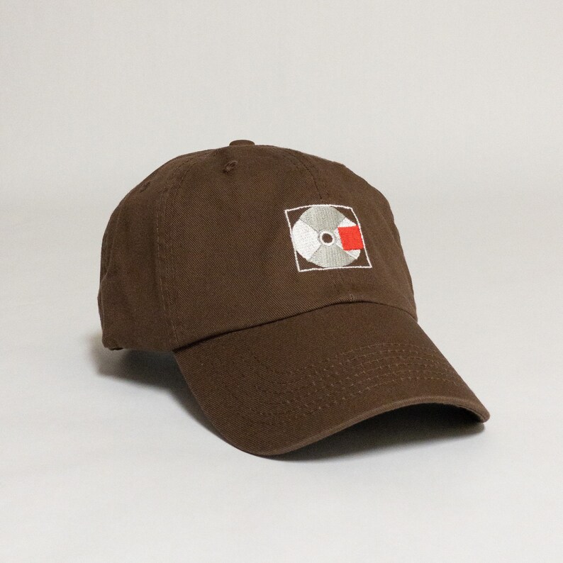 K&ouml;nnte beinhalten: Eine braune Baseballkappe mit einem wei&szlig;en und roten gestickten Logo einer Schallplatte mit einem roten Quadrat.