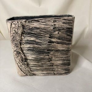 May include: A rectangular ceramic vase with a textured, wood-like appearance. The vase features a mix of beige and black tones, with horizontal and vertical lines creating a unique pattern. The interior is dark, and the vase has a slightly irregular shape.