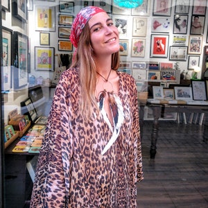 Puede incluir: Una mujer con un vestido con estampado de leopardo y un pañuelo rojo. El vestido tiene cuello en V y mangas largas. Sonríe frente a un escaparate lleno de arte enmarcado.