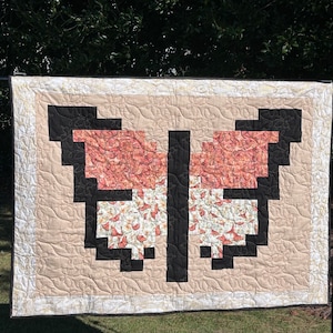 May include: A beige quilt with a black and white butterfly design. The butterfly is made up of smaller squares, each with a different floral pattern. The quilt is hanging on a fence.