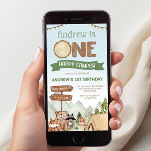 May include: A smartphone displays a birthday invitation with a camping theme. The invitation reads "Andrew is ONE Happy Camper" with woodland illustrations. The date and time are also included.
