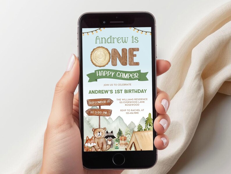 May include: A smartphone displays a birthday invitation with a camping theme. The invitation reads "Andrew is ONE Happy Camper" with woodland illustrations. The date and time are also included.