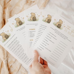 May include: Four baby shower game cards with Winnie the Pooh illustrations. The cards have gold and black text, with games like "The Price is Right" and "Get to Know the Other Shower Goer". The cards are on a cream-colored surface.